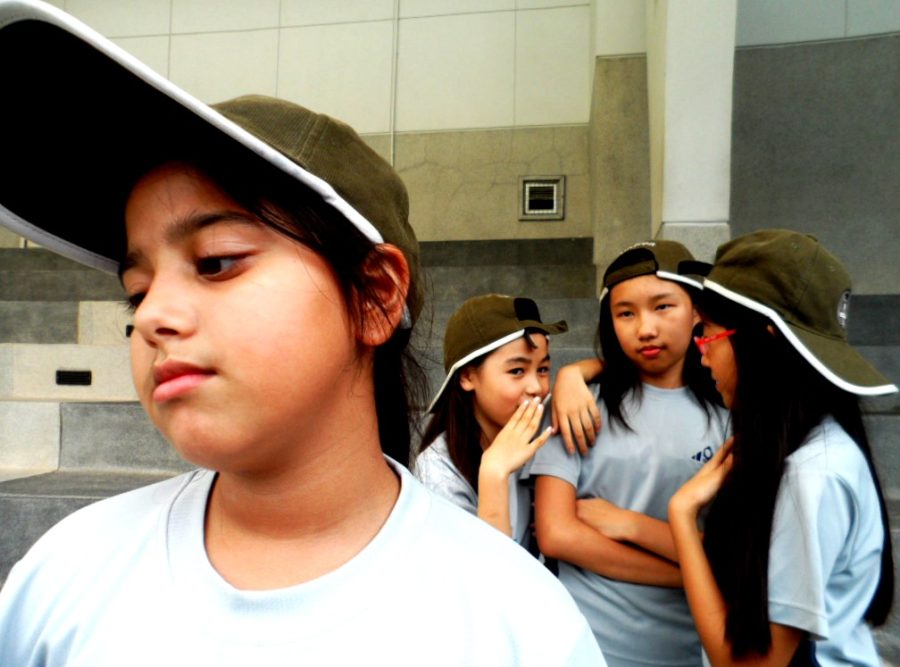 Three young girls whisper and verbally bully the girl with the cap. 