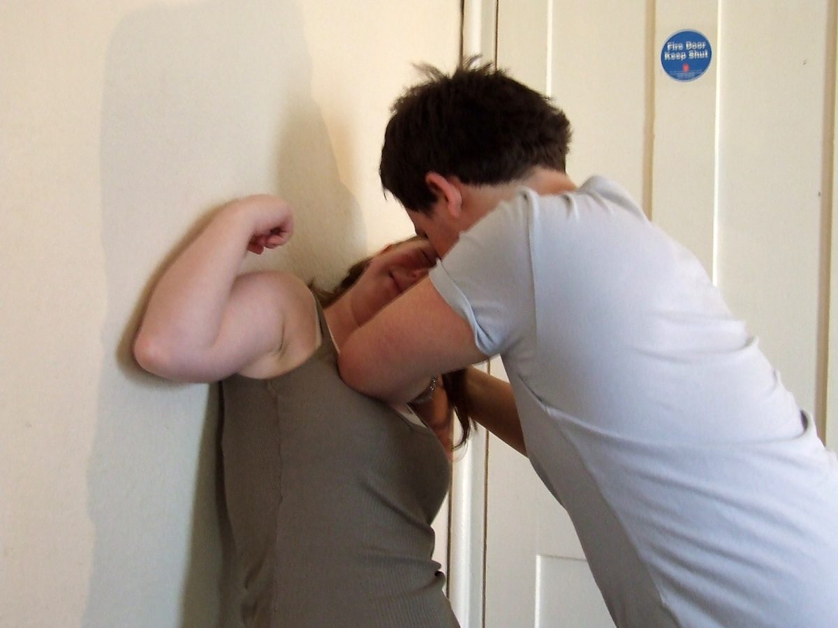 Man pushing a womans head into the wall with his elbow. 