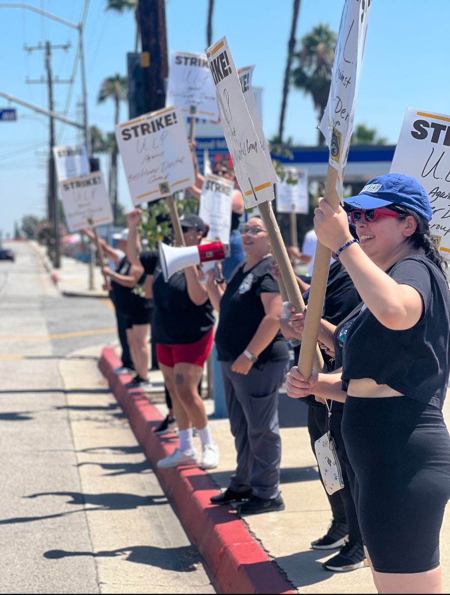 A row of people protesting with signs on the sidewalk of Rosecrans Ave.